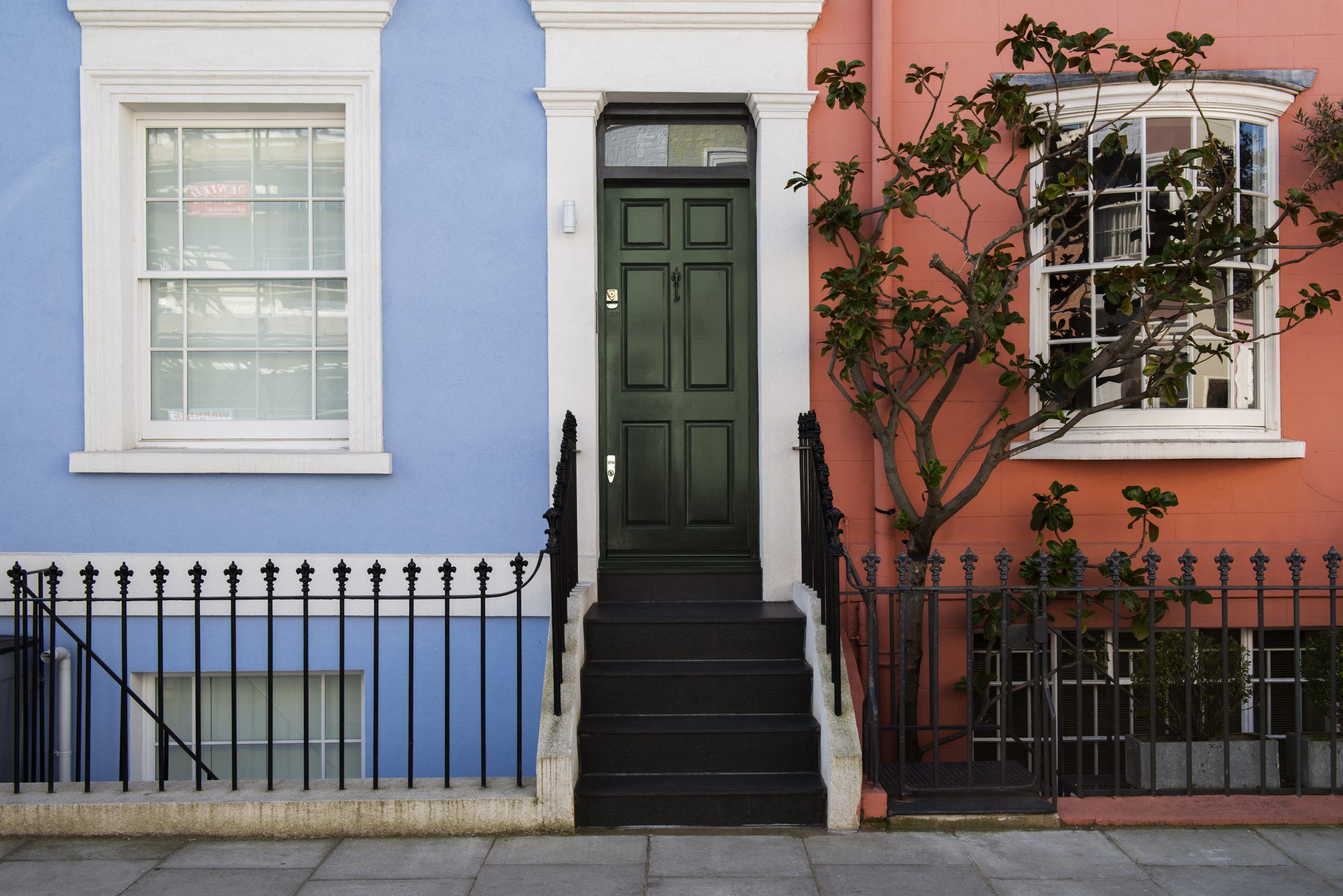A black front door with white trim and black steps on a blue wall with a white window above it, next to a pink wall with a white window and a tree, all separated by a black wrought iron fence. This is a typical example of the properties managed by Beacons Way Property Management.